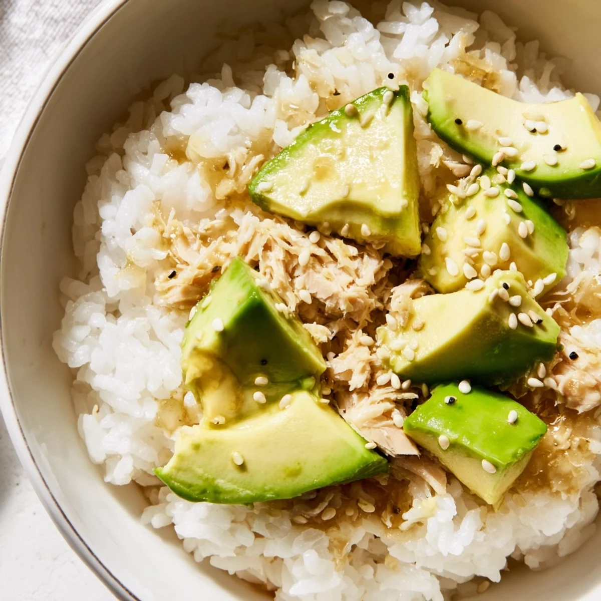 A close-up of a vibrant tuna avocado rice bowl, with fluffy jasmine rice topped with diced tuna, creamy avocado, crisp cucumber, scallions, and toasted sesame seeds.  